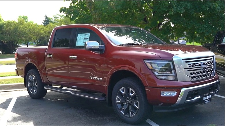 A red Nissan Titan pickup truck parked at a dealership lot, with a window sticker visible on the driver's side and a Walser Automotive Group license plate frame on the front.