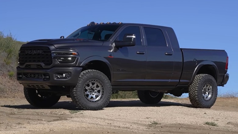 A lifted black 2025 Ram 2500 Cummins pickup truck with off-road tires is parked on a dirt path with a small forested background.