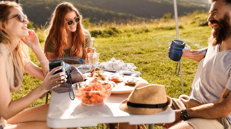Three people enjoying picnic outdoors holding portable speakers.