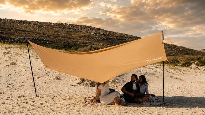 A family sitting beneath tarp camp in sandy surface.