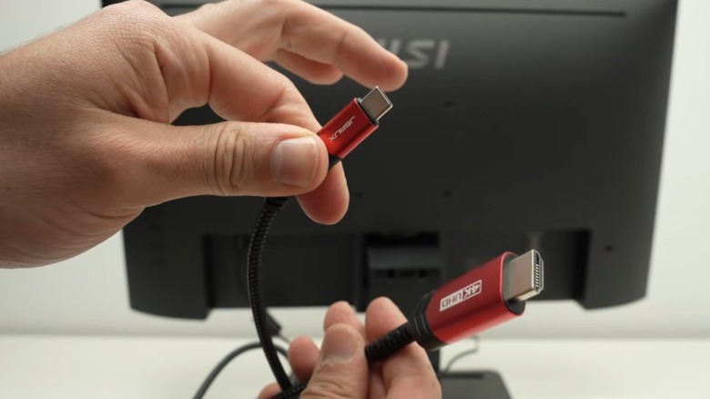 Hands holding both ends of a USB-C to HDMI video cable with computer monitor in the background