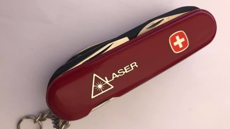 A red Swiss Army-style knife with a keyring, labeled "LASER" alongside a laser hazard symbol and a white cross emblem on the handle.