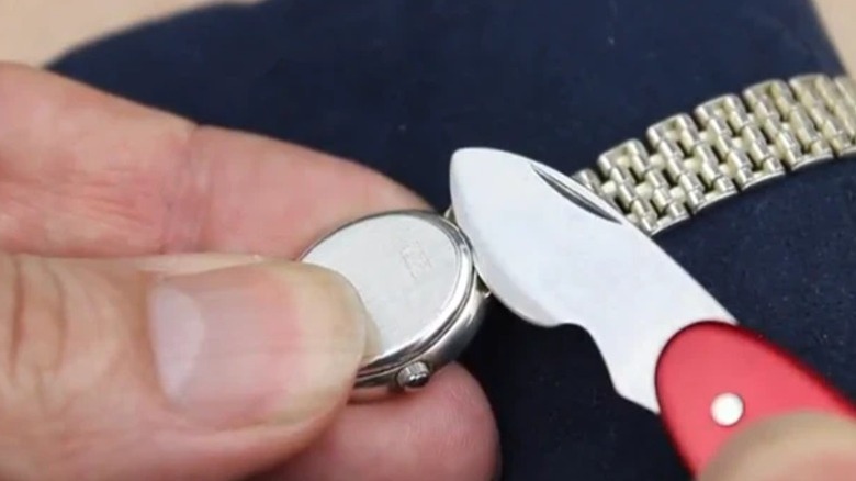 A close-up of a person using the watch case opener blade of a red Swiss Army knife to carefully pry open the back of a wristwatch.