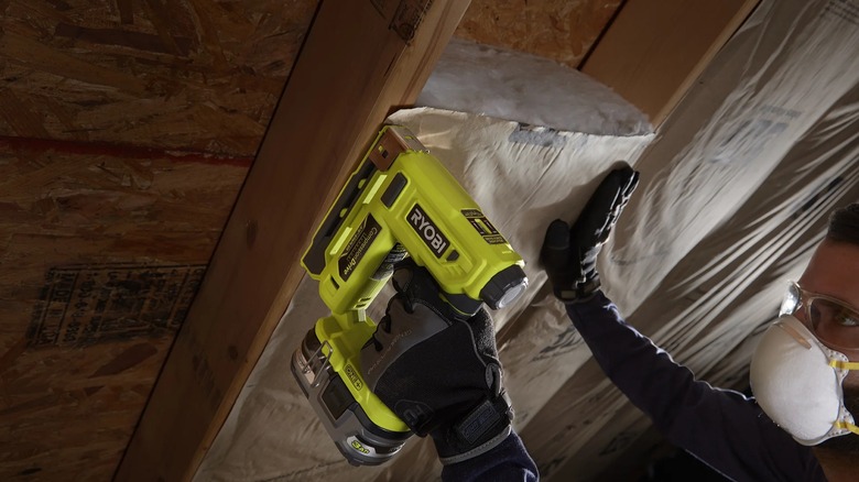 A worker wearing gloves and a face mask securing insulation with a Ryobi stapler