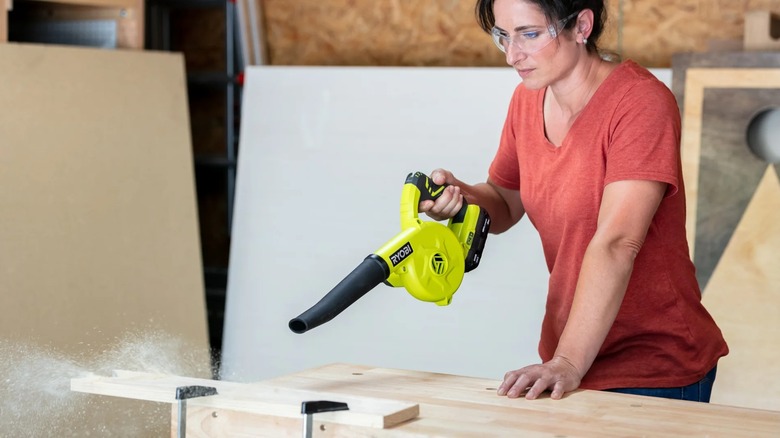 A person wearing safety glasses blowing sawdust off a workpiece with a Ryobi blower