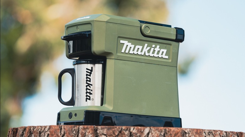 A green Makita Outdoor Adventure 18V LXT cordless coffee maker sitting on a stump.