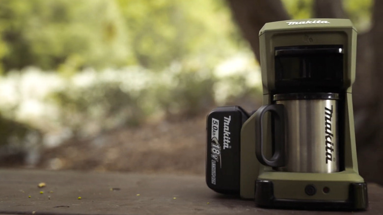 A Makita Outdoor Adventure 18V LXT cordless coffee maker and stainless steel Makita mug on a wooden picnic table.