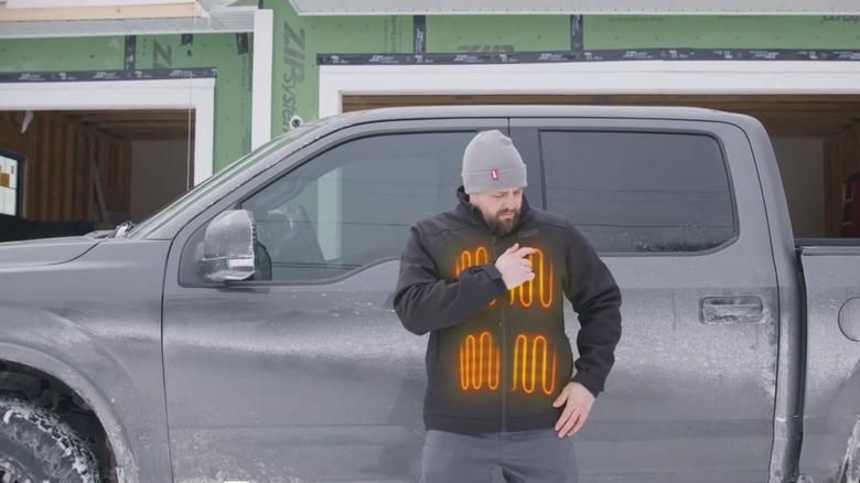 A man wearing a heated Milwaukee jacket with glowing orange heat zones visible on the chest and abdomen stands outside beside a gray pickup truck.
