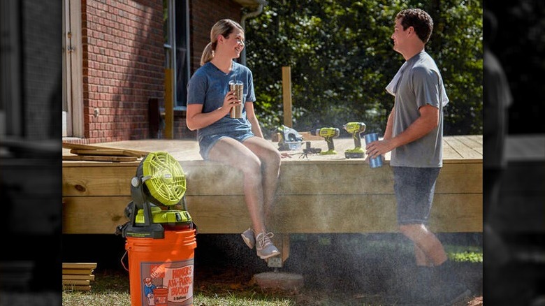 A Ryobi 18V ONE+ bucket-top misting fan sprays a cooling mist from a bright orange bucket while two people relax with drinks on a wooden deck.
