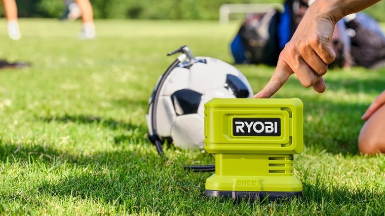 A close-up of a person pressing the power button on a bright green Ryobi ONE+ inflator set on a grassy soccer field.