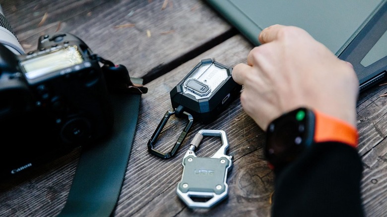 UAG Monarch carabiner with AirPods and a camera on a wooden table