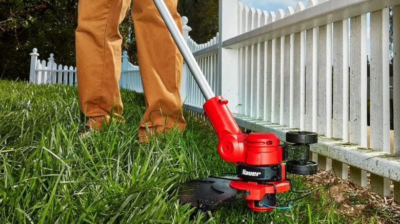 Person using a Bauer string trimmer to cut grass next to a white picket fence