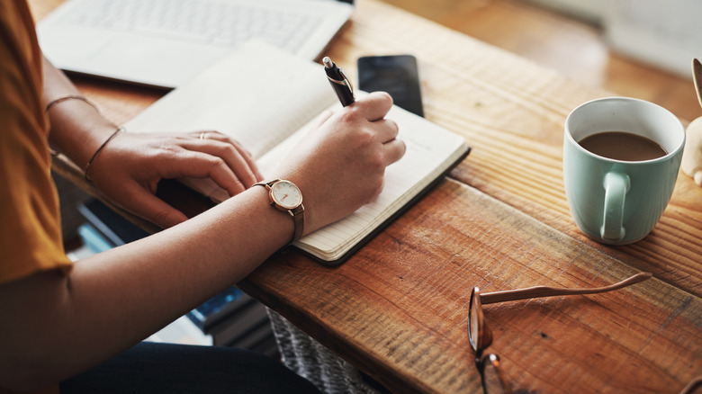 A person writing in a notebook at a table with a computer and a cup of coffee