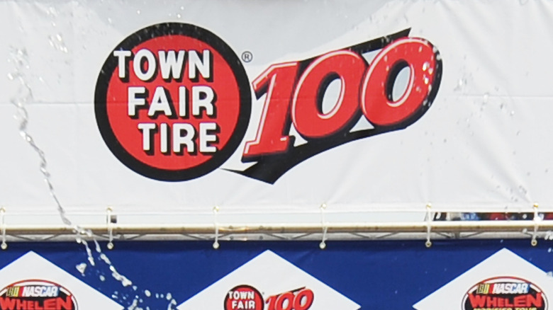 The Town Fair Tire logo as part of the Town Fair Tire 100 sporting event printed on a flex billboard on a white backdrop.