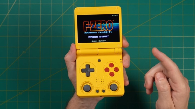 Hand holding a yellow Anbernic RG 34XXSP handheld console above a green backdrop