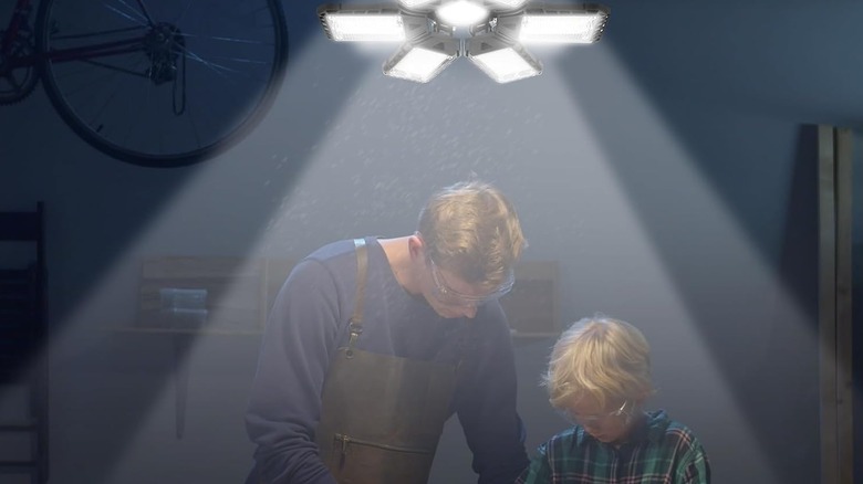 An adult and a child work on a project in a garage under a large LED fixture.