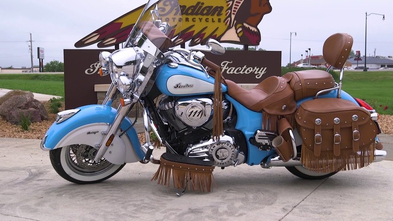 Blue and white Indian motorcycle with leather saddle bags, seat, and accessories parked on concrete in front of an Indian Motorcycle Factory sign
