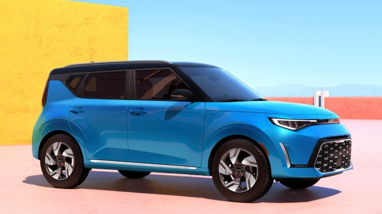 Blue Kia Soul parked on a pink surface with a yellow wall and blue sky in the background.