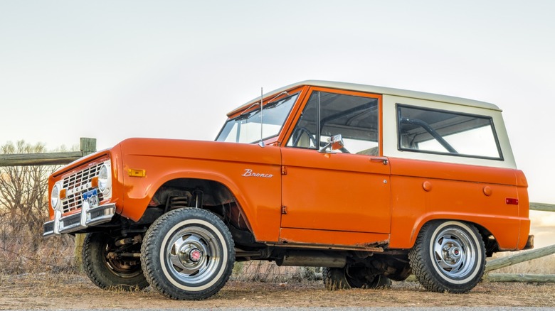 a first-generation Ford Bronco in orange color parked near a farm fence