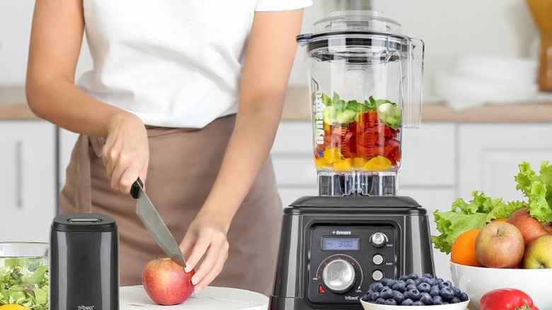 Tribest Dynapro blender on a table with bowls of fruit and a person chopping fruit in the background