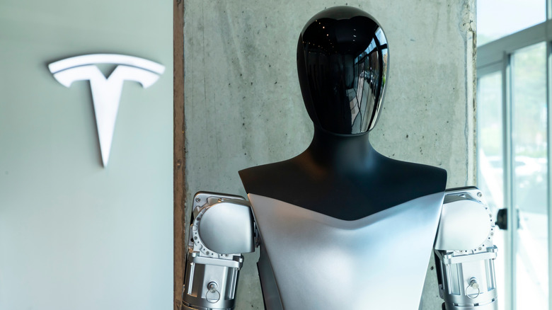 The Tesla Optimus robot standing in front of the Tesla logo