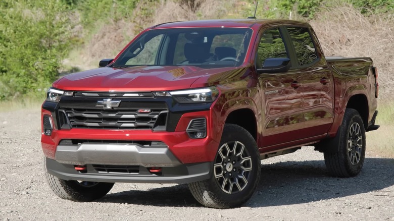A deep red Chevrolet Colorado truck on a loose gravel road with green brush filling the background.