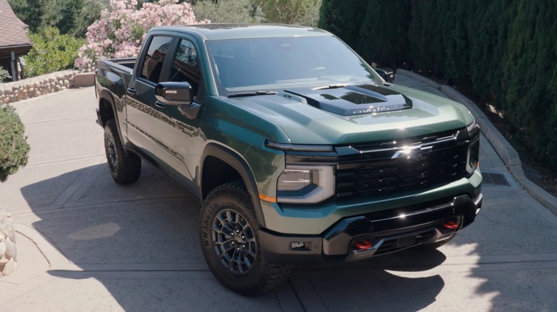 A deep forest green 2027 Chevrolet Silverado parked at the top of a steep residential driveway surrounded by tall cypress trees and blooming pink flowers.