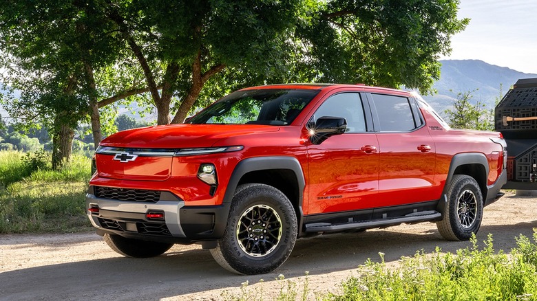 A red 2026 Chevrolet Silverado EV Trail Boss crew cab sitted on a dirt road beside a tree and a mountain lake, hooked up to a black-and-orange overland trailer.