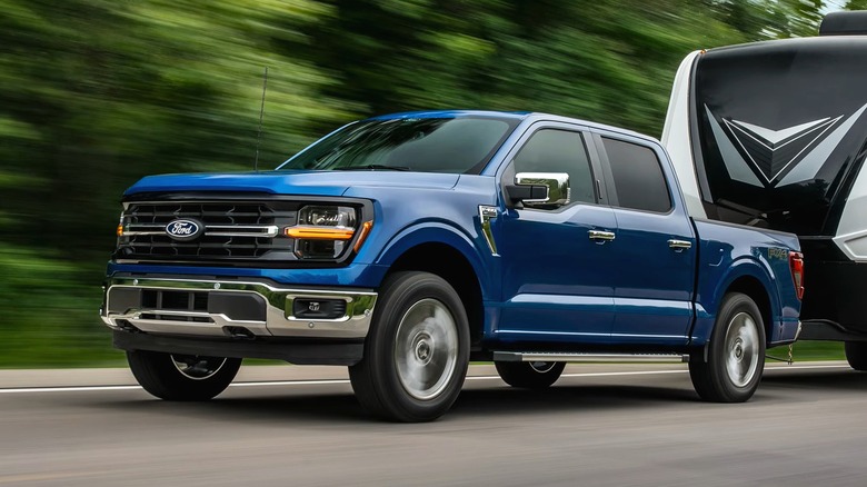 A blue 2026 Ford F-150 crew cab pulling a large black-and-white travel trailer down a tree-lined highway at speed.