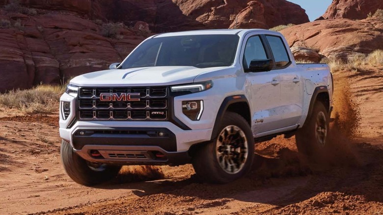 A white 2026 GMC Canyon AT4 kicking up a plume of red desert dust as it carves through a sandy wash surrounded by sandstone formations.