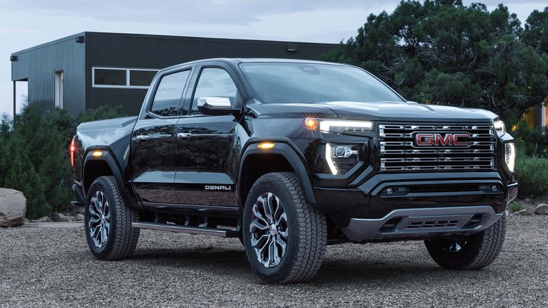 A gloss black 2026 GMC Canyon Denali crew cab sitting on a gravel lot at dusk in front of a modern dark building.
