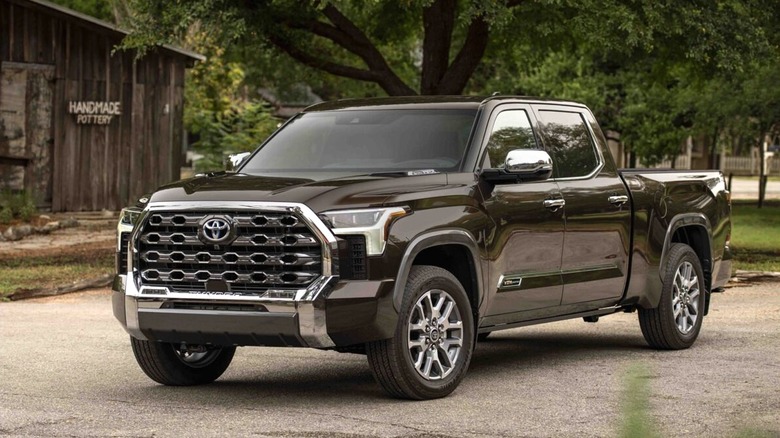 A dark brown 2024 Toyota Tundra 1794 Edition Smoked Mesquite 054 truck on a quiet road in front of a weathered timber barn and a large shade tree.