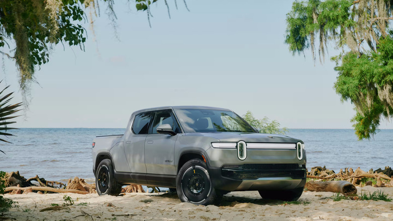 A gray first-gen Rivian R1T standing on a beach, trees on the sides and water in the background.