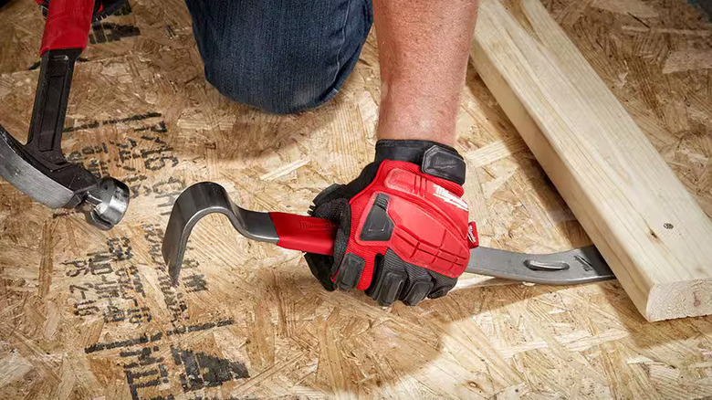 A gloved worker using a Milwaukee pry bar and a hammer to lever up a nailed-down 2x4
