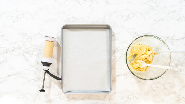 Old-fashioned cookie press next to a baking sheet and bowl of dough.