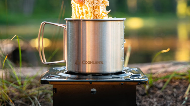 Coghlan's cooking stove with a Coghlan's cooking cup on top as someone puts in noodles