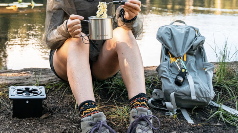 A person sitting near a Coghlan's portable folding camp stove eating noodles out of a Coghlan's cooking mug