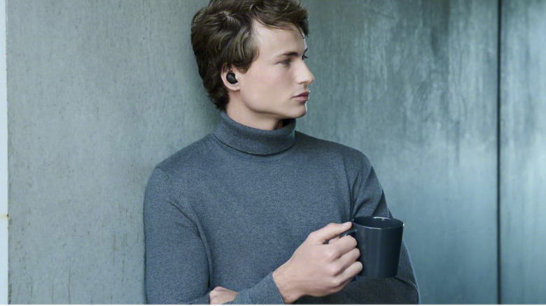 A person wearing Sony XM6 earbuds leaning against the wall holding a cup.