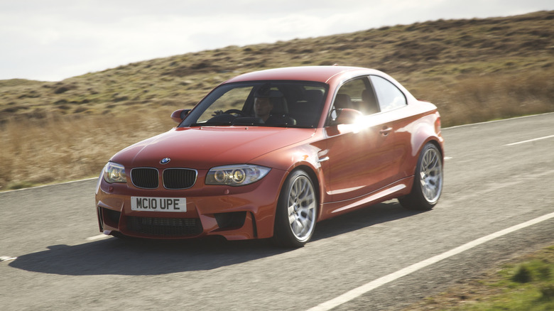 An orange BMW 1 Series M Coupe driving on an empty road.