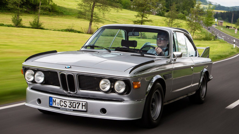 A silver BMW E9 CSL driving with trees in the background.