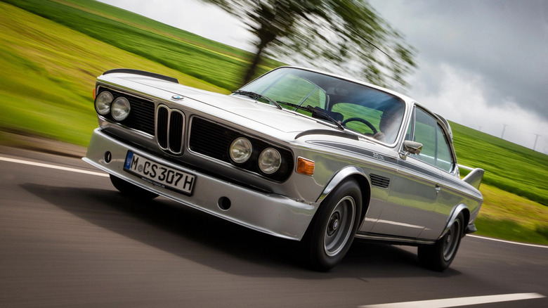 A silver BMW E9 CSL driving down a road.