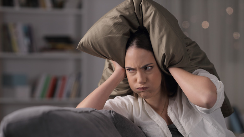 A woman with a frustrated expression on her face covering her ears with a pillow