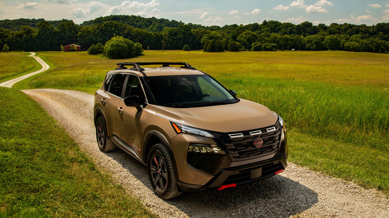 A brown 2026 Nissan Rogue on a dirt road surrounded by fields and trees on a sunny day