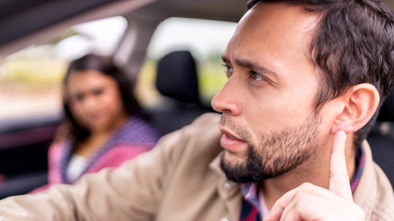 A man with a woman in a car pointing to his ear to indicate he's listening to something
