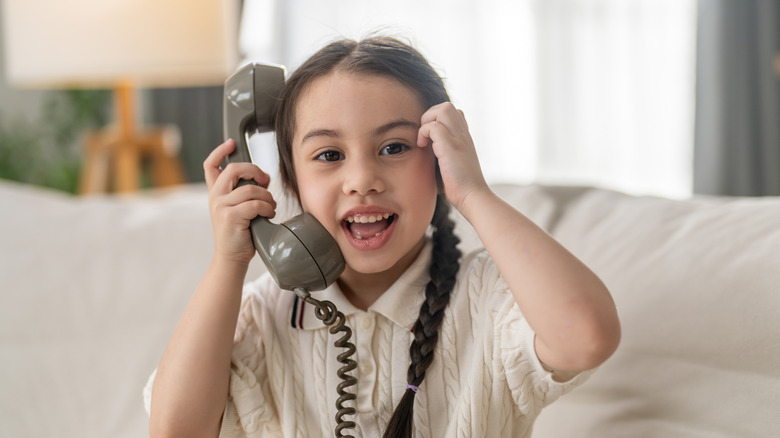 A young girl sitting on a white sofa holding up an old-fashioned phone receiver