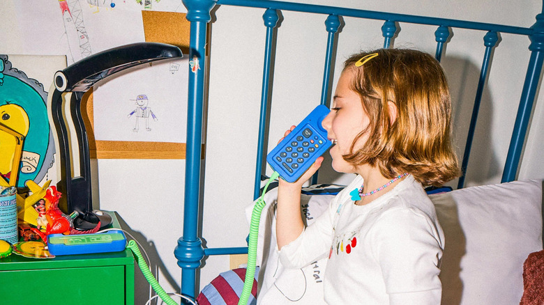A young girl sitting on a blue bed using a blue and green Tin Can phone in a cluttered bedroom