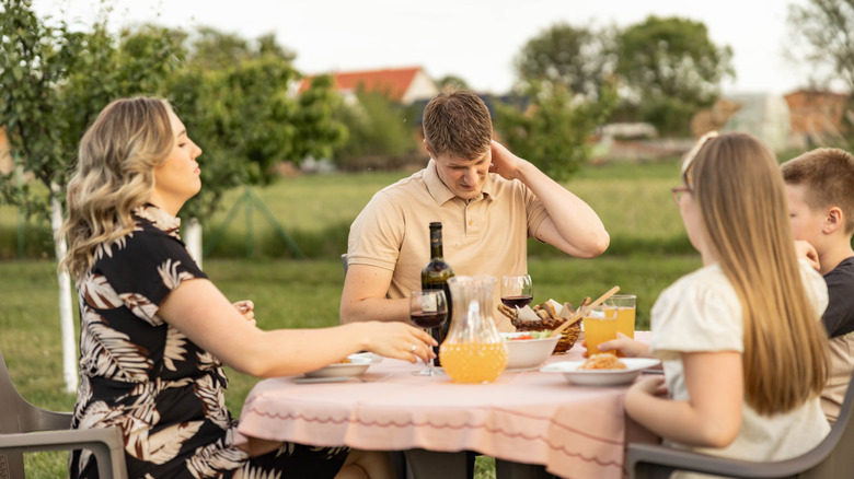 Family eating at a table outdoors, with one man scratching his neck