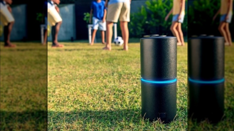 Grassy yard with people playing soccer, with a black cylindrical device in the foreground