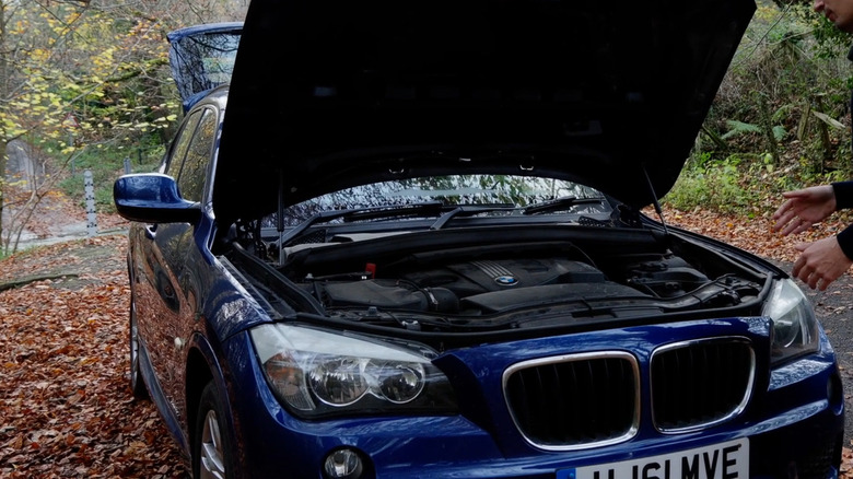 A man's pair of hands reaching towards the open engine bay of a diesel BMW X1 from the late 2000s.
