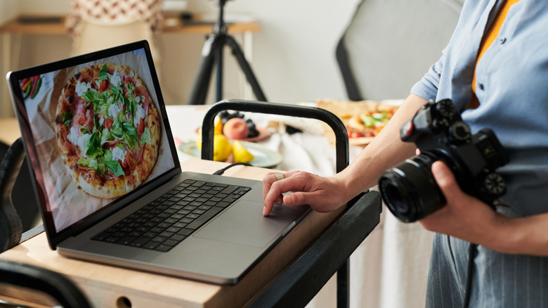 A person holding a digital camera while using laptop with a photo of a pizza on the screen